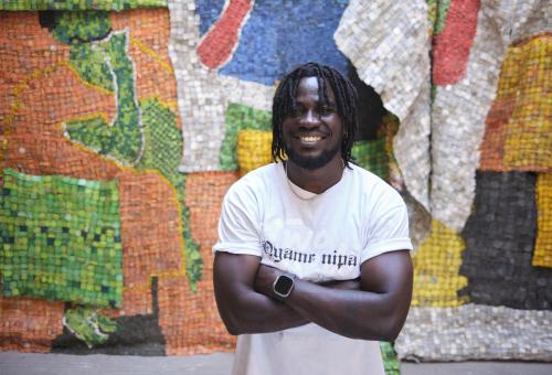 Serge Attukwei Clottey smiling and stood with his arms crossed in front of a colourful artwork